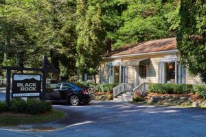 black rock tile and design showroom nestled in a wooded setting. a black car sits parked outside this luxury bath and tile design business. the building features a quaint, cottage-like design.