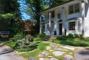 elegant white house exterior with stone walkway and lush landscaping. sign reads "black rock design studio cabinetry".