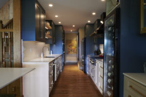 a modern, mountain contemporary kitchen featuring dark blue cabinets, white countertops, and wood floors. a wine fridge stands to the right, with a colorful painting visible at the end of the galley kitchen.